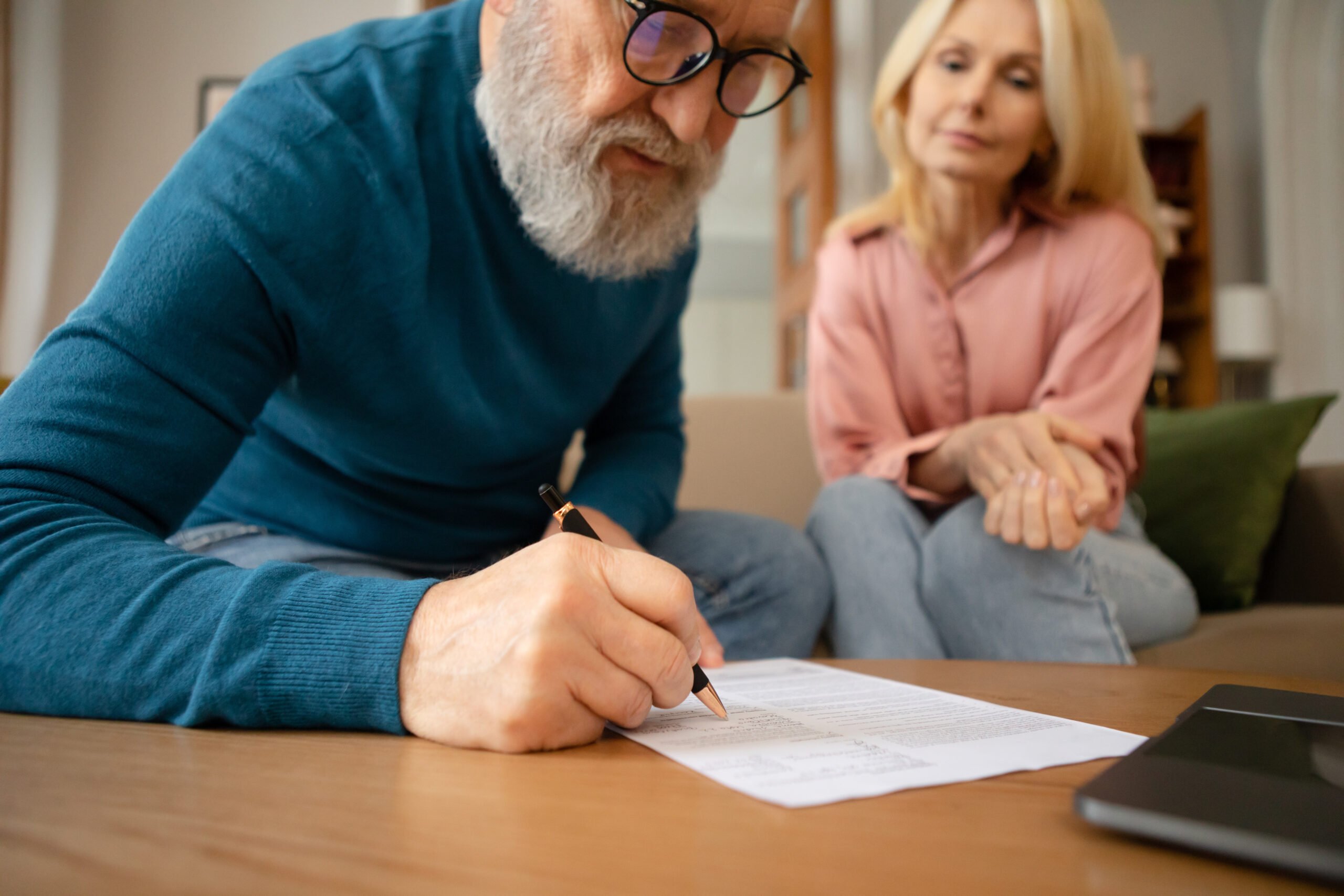 Senior Couple Filling In Form Signing Papers Sitting On Sofa Near Table Indoors. Paperwork Concept. Elderly Man Writing His Will In Notary Office. Cropped Shot, Selective Focus
