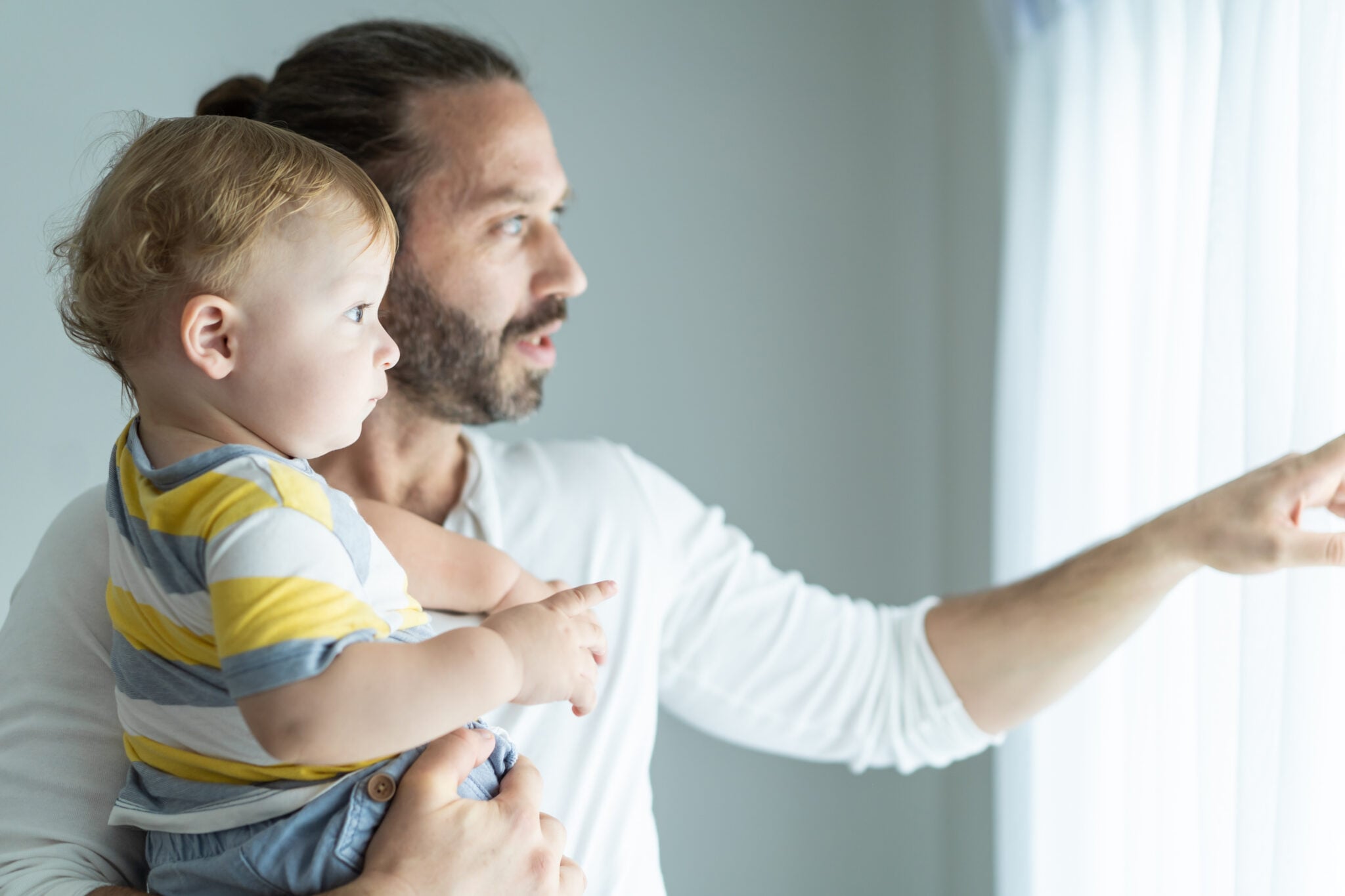 Caucasian loving father holding young baby boy child in living room. Happy family, attractive caring daddy carry his toddler son on hands and pointing to window in house. Activity relationship at home