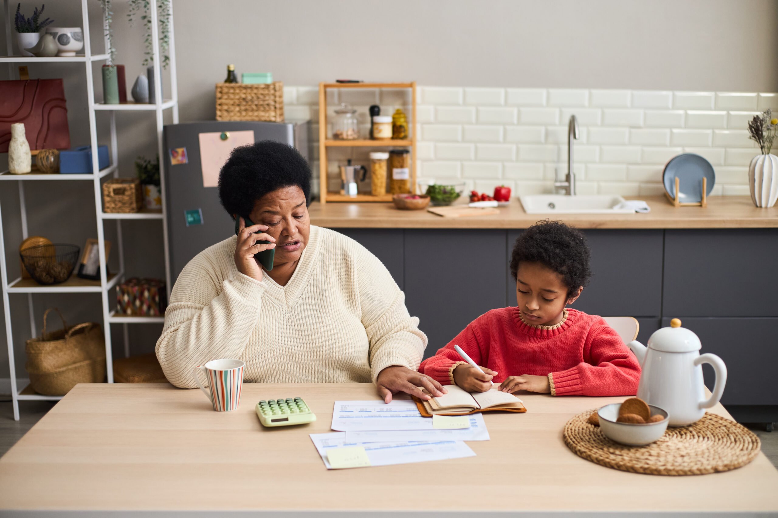 Middle aged Black woman talking on smartphone while sitting at kitchen table with young Black boy doing homework, documents and calculator on table, modern kitchen background