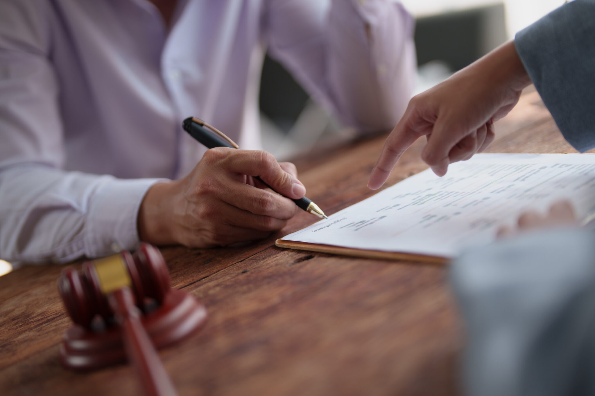 Person's hand signing a legal document on a wooden table, with another person pointing to a specific line, representing contract signing, agreement, or legal proceedings
